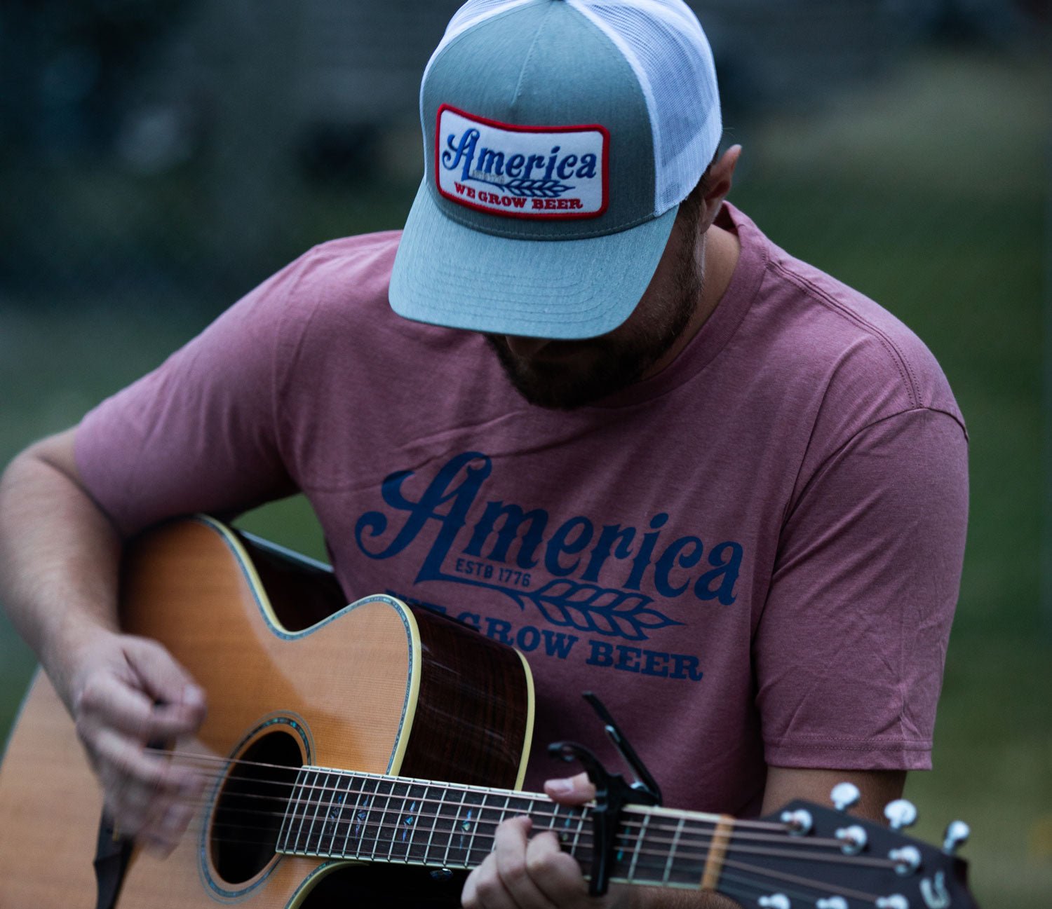 A person wearing a cap and a pink "America, We Grow Beer" shirt is playing an acoustic guitar outdoors. The image focuses on the individual's upper body and guitar, with a blurred background.