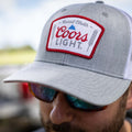 Close-up of a person wearing the Rc X CL Hat by Rural Cloth, featuring a grey baseball cap with a prominent Coors Light logo. The snapback closure ensures a perfect fit, while the top prominently displays 