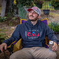 A man sitting in a purple and yellow outdoor chair is holding a can of Coors Light® American beer. He is wearing a grey hoodie with the Coors Light® logo and a We Grow Coors Light Hat by Rural Cloth, which features grey and white colors with red lettering. There are plants and greenery in the background.