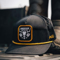 A black baseball cap from Rural Cloth’s Skull & Roses Hat collection is resting on a surface. The cap features a yellow rope detail along the brim and a patch with the text 