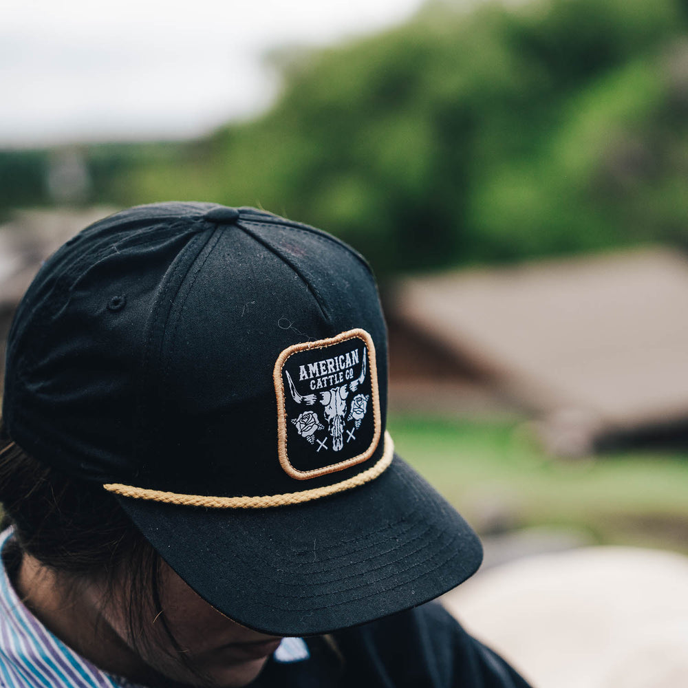 Person wearing the Skull & Roses Hat by Rural Cloth, which features an adjustable snapback and a patch that reads "American Cattle" with roses and crossed arrows. The background showcases blurred greenery and a brown structure, suggesting an outdoor setting.