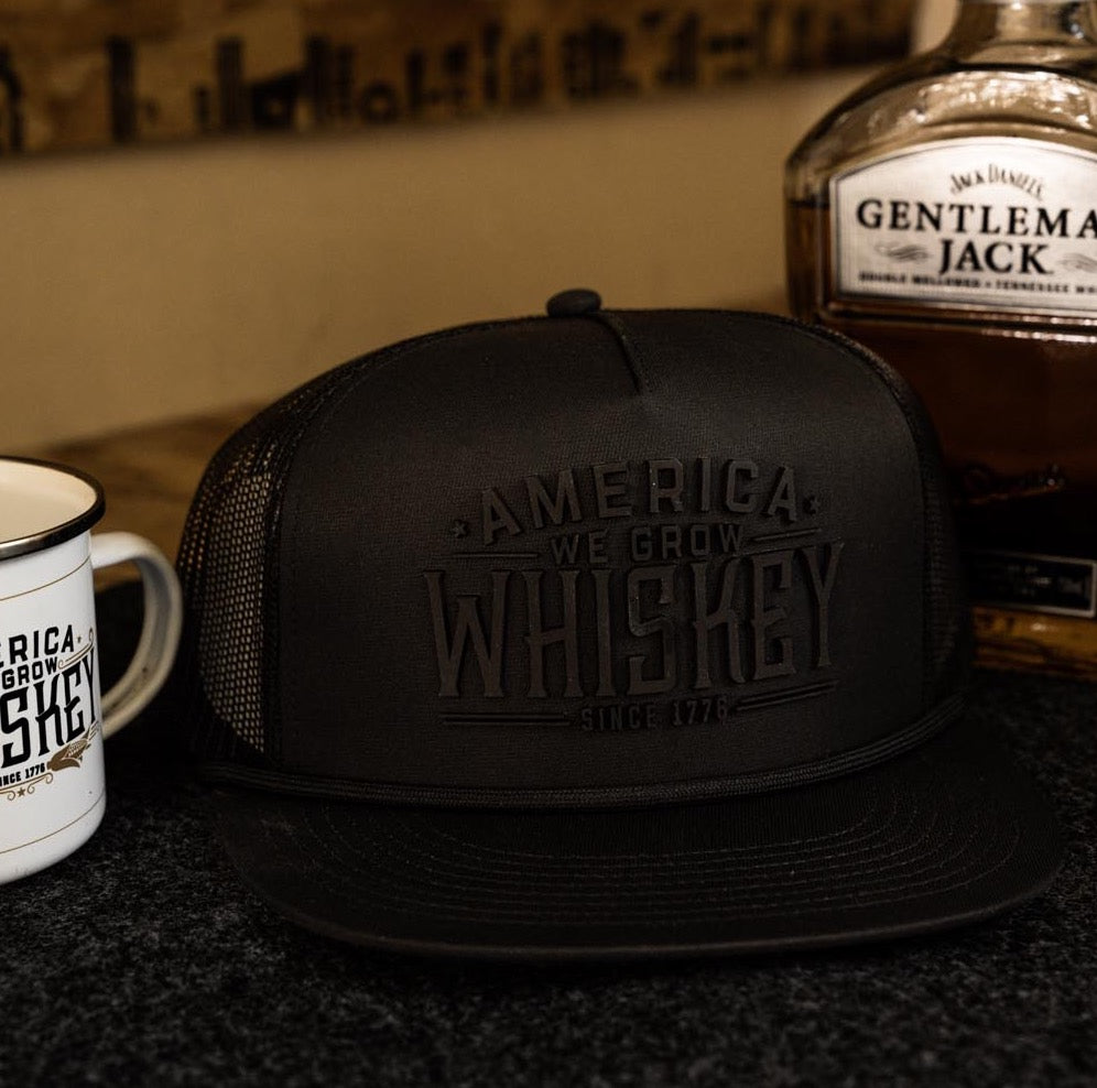 A cap with a snapback closure and a mug branded with "We Grow Whiskey - Flat Silicon (WGWS-F01)" text are placed on a flat surface. A bottle of Gentleman Jack Tennessee whiskey sits next to them. The background features a blurred image of what appears to be a wooden wall with various inscriptions, paying homage to corn farmers.