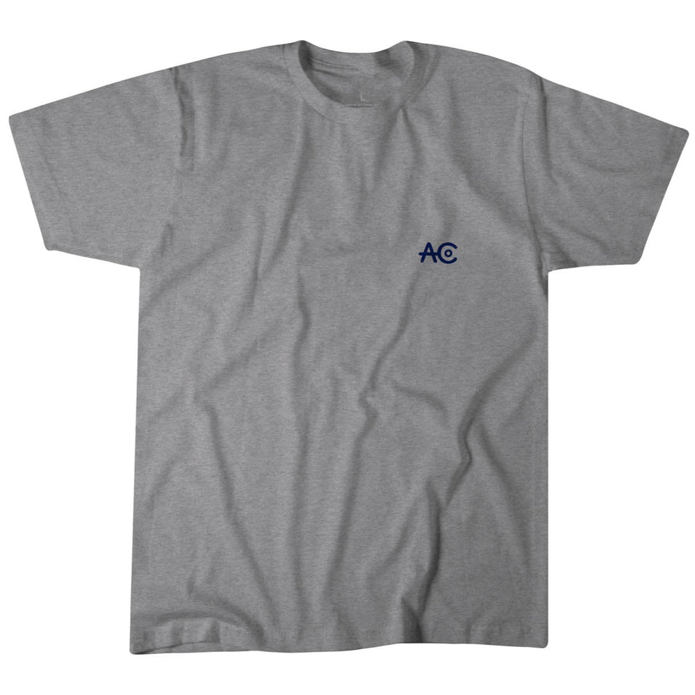 The AC Flag Tee by Rural Cloth is a gray T-shirt featuring a small navy blue "AC" logo on the left chest, embodying classic American ranching style. The soft fabric shirt has short sleeves and a round neckline, displayed flat against a white background.