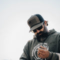 A person with a beard wearing sunglasses and a breathable ACC Flag Hat by Rural Cloth, featuring an American flag design, is looking down while holding an object. The person is dressed in a hoodie that bears the logo and text for 