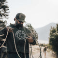 A bearded man wearing a hoodie and a Rural Cloth ACC Flag Hat, featuring breathable mesh and an American Cattle graphic, is handling a coiled rope outdoors. He is also sporting sunglasses. In the background, trees and mountains can be seen with someone walking away in the distance, capturing the essence of ranching life.