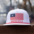 A white Rural Cloth ACC Flag Hat with a red brim sits on a brick surface, displaying the American Cattle Co design. It prominently features an American flag with a bull skull and stars on the left side. Embrace the ranching lifestyle with this breathable mesh snapback, adorned with 