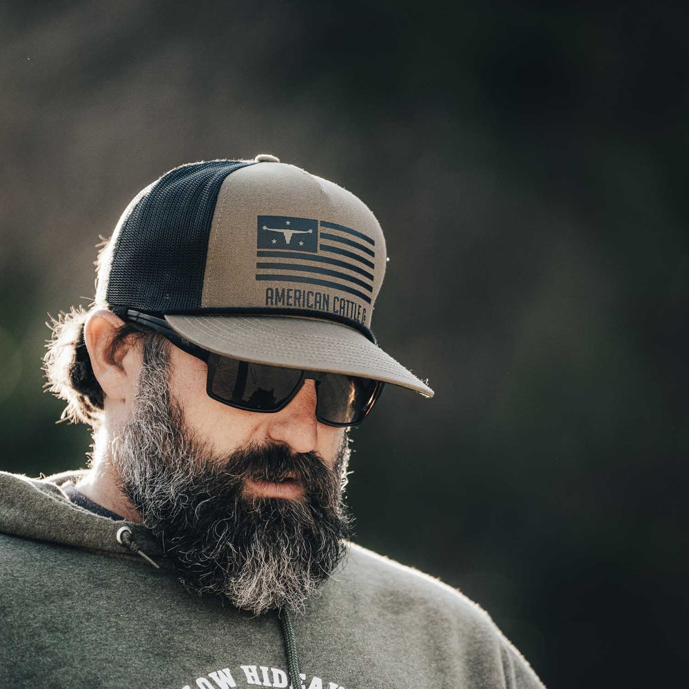 A bearded man is wearing sunglasses, a hoodie, and the ACC Flag Hat by Rural Cloth, featuring an "American Cattle" graphic emblematic of his ranching lifestyle. The breathable mesh snapback hat sits comfortably on his head as he glances downward, with a blurred, dark background out of focus behind him.