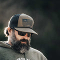 A bearded man is wearing sunglasses, a hoodie, and the ACC Flag Hat by Rural Cloth, featuring an 