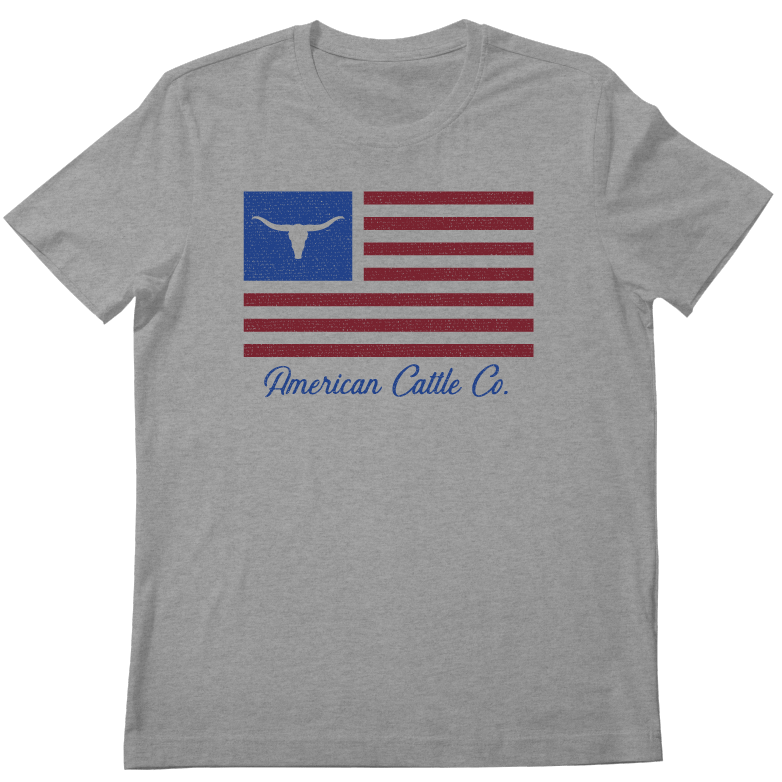 The ACC Flag Tee by Rural Cloth is a gray T-shirt featuring an American flag design with a blue rectangle and a white bull skull silhouette in place of the stars. Below the flag, it reads "American Cattle Co." in blue text, capturing the rugged spirit of American cattle ranching.