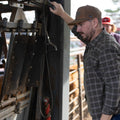 A man wearing an ACC Waxed Canvas hat from Rural Cloth and a plaid shirt operates a piece of machinery at a cattle farm. Another person, with an embroidered design on their red cap, stands in the background near cattle in a fenced area. The scene appears to be outdoors, with farming equipment and livestock visible.