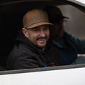 A man with a beard is sitting in the driver's seat of a car, wearing the ACC Waxed Canvas hat from Rural Cloth, which features an embroidered design and the word 