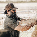 A bearded man wearing sunglasses and an ACC Waxed Canvas hat from Rural Cloth is spreading straw. He has on a grey t-shirt and is set against an outdoor, rural backdrop.