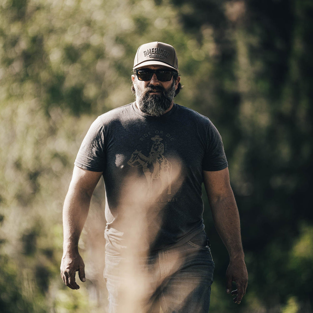 A bearded man wearing sunglasses and a waxed canvas Rural Cloth ACC hat with a snapback closure, along with a graphic t-shirt, walks outdoors surrounded by greenery. He has a serious expression and his hands are down by his sides.