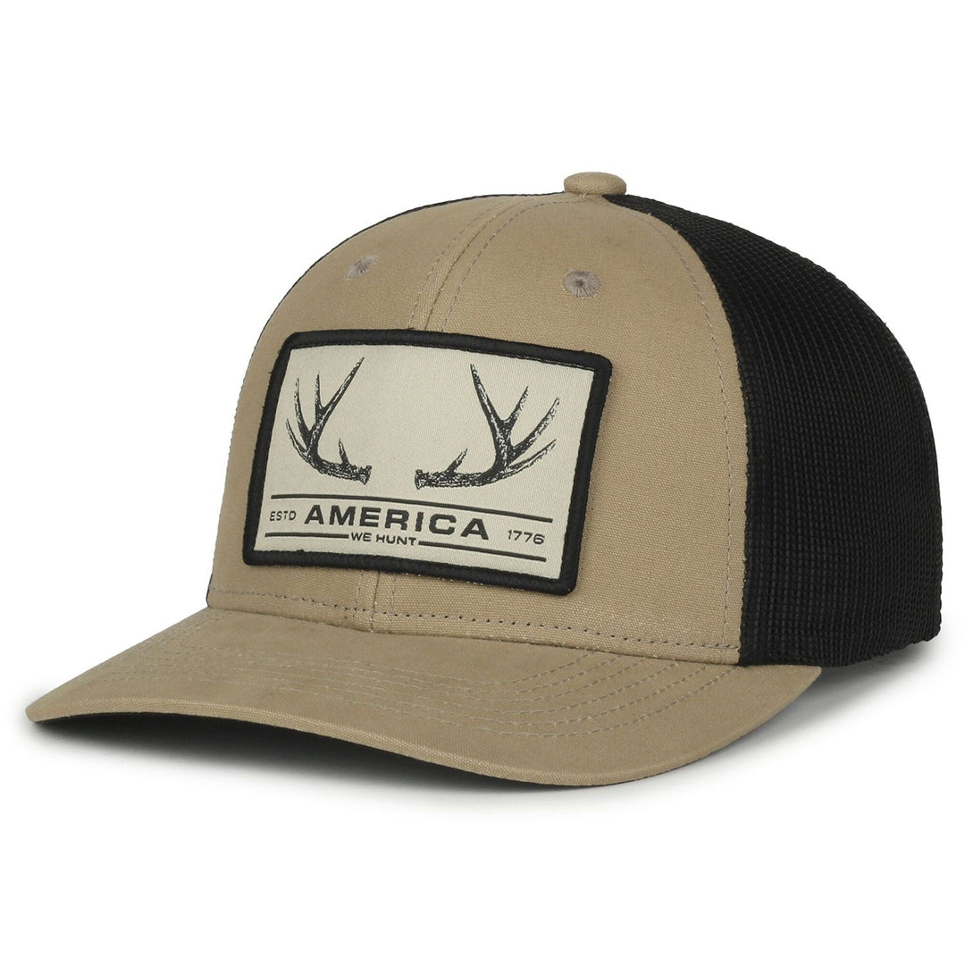 The America We Hunt Deer Hat from Rural Cloth is a tan and black mesh baseball cap featuring a snapback closure and gray eyelets. The cap has a curved brim and a front rectangular patch displaying antlers with the text "AMERICA WE HUNT" above "ESTD 1776.