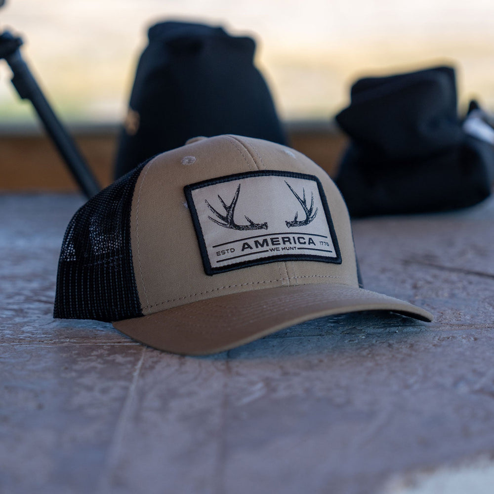 A light brown and black trucker hat with a mesh back and snapback closure rests on a weathered surface. The hat, branded as the "America We Hunt Deer Hat" by Rural Cloth, features a patch with antler graphics and the text "America," along with smaller text reading "ESTD 1776" and "LIVE HUNT." Blurred background items are visible.