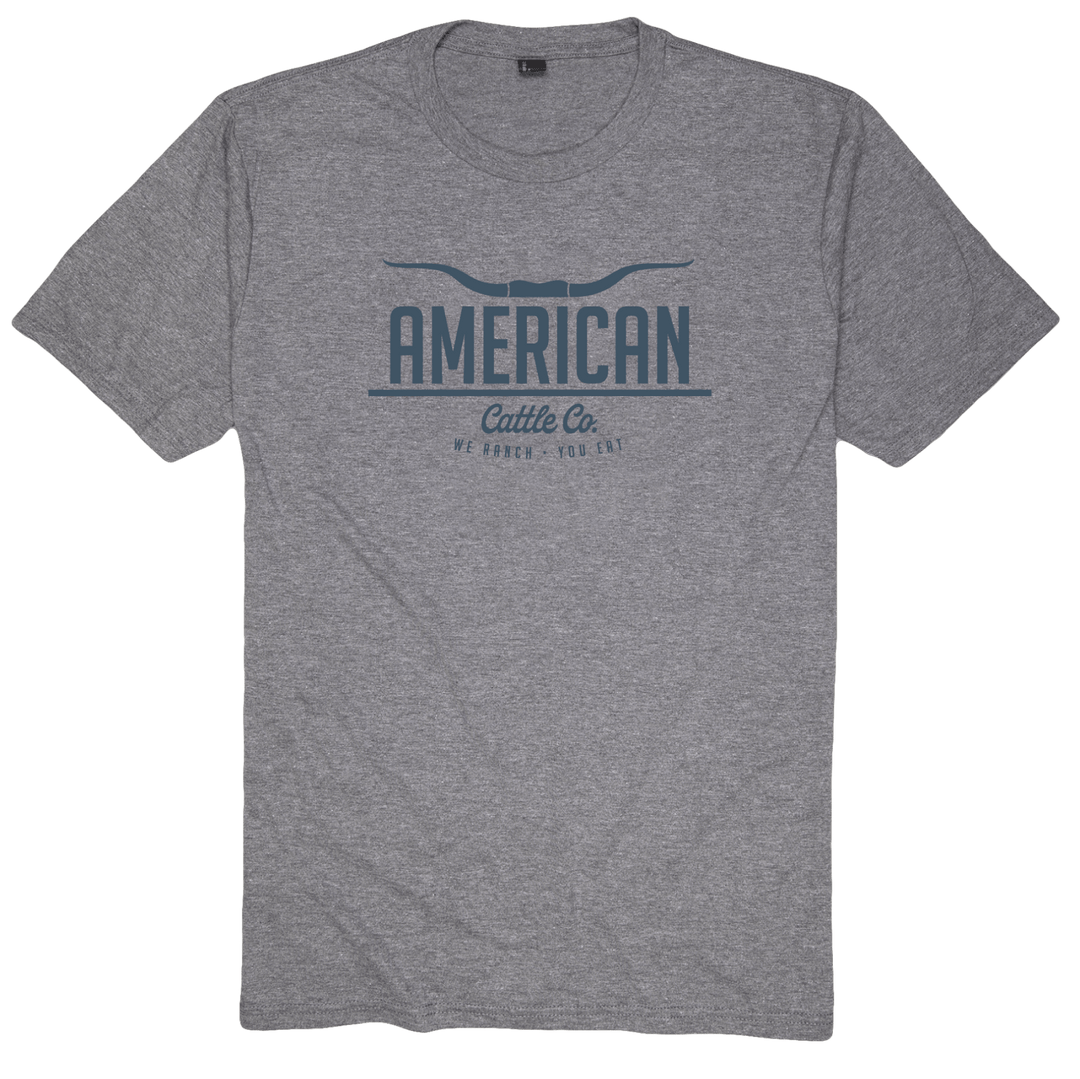 The American Cattle Co Tee by Rural Cloth features a heather gray fabric adorned with the bold, capitalized text "AMERICAN Cattle Co." along with a striking longhorn graphic above it. Below, in an elegant font, the phrase "WE RANCH. YOU EAT." adds a touch of charm, celebrating the true American spirit of farming and ranching.