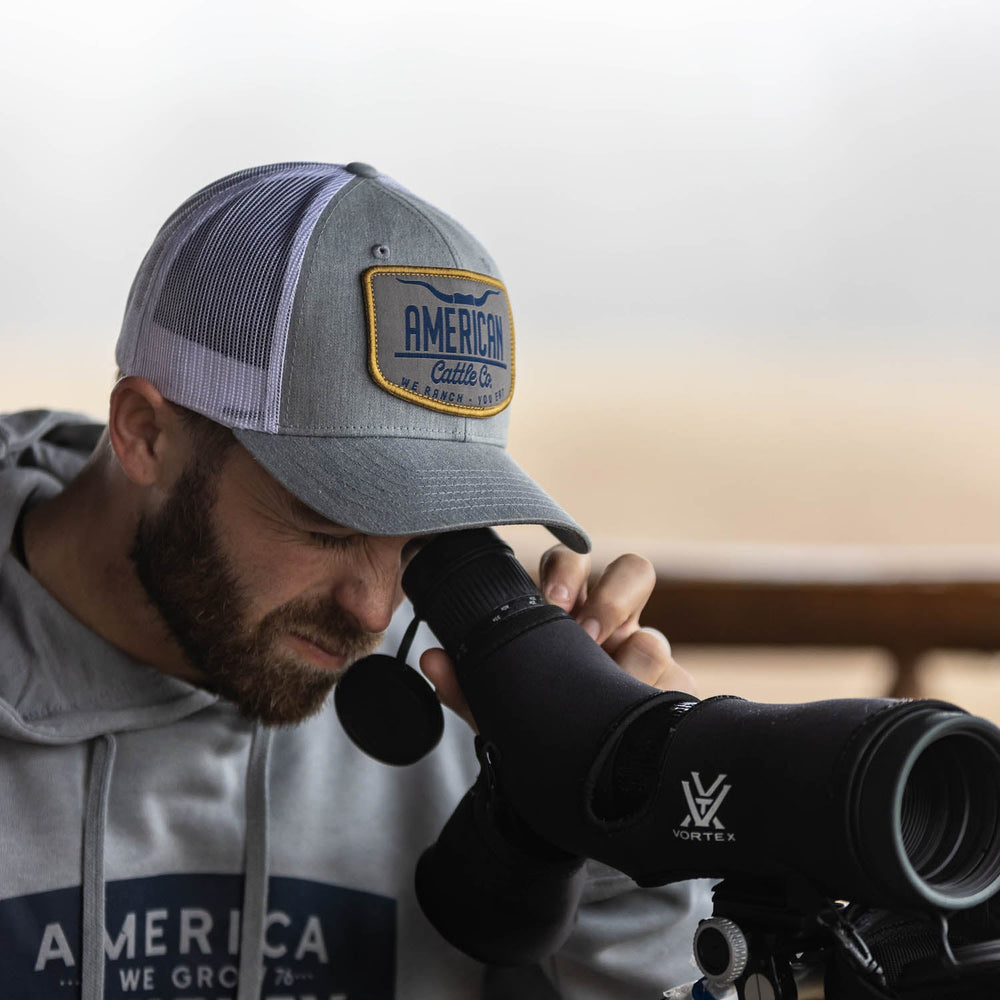 A man is observing through a spotting scope, wearing the Rural Cloth American Cattle Co Hat in Gray with a matching gray hoodie. The backdrop suggests an outdoor setting, likely on a porch or deck, embodying the quintessential American farming and ranching lifestyle.