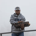 A man wearing a gray hoodie and an American Cattle Co Hat-Gray by Rural Cloth is carrying a large log. He has a beard and is sporting gloves, embodying the rugged American lifestyle. The foggy background hints at an outdoor setting, reminiscent of traditional farming and ranching activities.