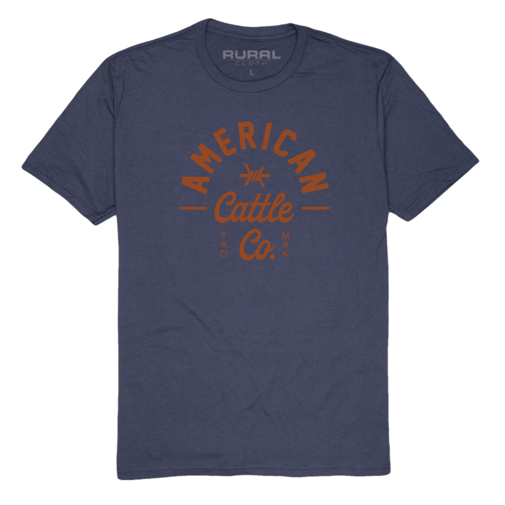 The Barb Tee from Rural Cloth is a blue t-shirt featuring the text "American Cattle Co." printed in orange on the front. The words "RURAL CLOTH" are printed on the inner neckline. Made from premium weight fabric, this resilient shirt boasts a simple design with short sleeves and a crew neck.
