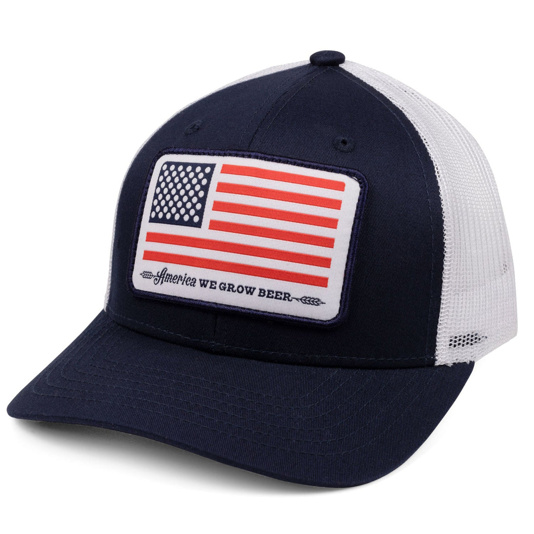 The Rural Cloth Bottle Cap Hat is a navy blue and white trucker cap adorned with an American flag patch on the front. Beneath the flag, the text reads, "America WE GROW BEER," flanked by a beer hop design on both sides. Celebrating American beer culture, this patriotic cap features a mesh back, curved brim, and adjustable snapback closure.