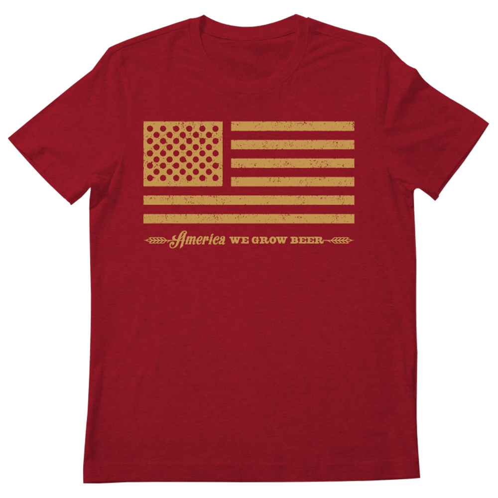 A red Bottle Cap Tee from Rural Cloth showcases a distressed yellow American flag design on the front. Beneath the flag, the text "America WE GROW BEER," flanked by small wheat graphics, makes it an ideal piece for patriotic celebrations and a perfect embodiment of American beer culture.