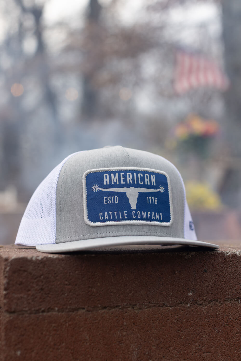 The Bull Spurs Hat-Flat by Rural Cloth is a stylish gray and white trucker hat with a flat bill, adorned with a blue and white patch on the front. The patch displays "American Cattle Company," "ESTD 1776," along with an image of a cow's head. The hat rests on a brown brick surface, with blurred outdoor scenery in the backdrop.