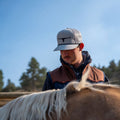 A person wearing a Rural Cloth Bull Spurs Icon Hat and a brown and black jacket stands closely beside a horse. The person's face is partially obscured by the brim of the hat. Trees and a blue sky are visible in the background.