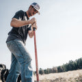 A man wearing a Bull Spurs Icon Hat by Rural Cloth and a dark T-shirt is using a long-handled tool to dig or break ground in an open field. A black dog stands nearby, with trees and a clear sky in the background.