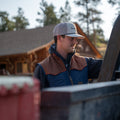 A man wearing a blue and brown vest with a Rural Cloth Bull Spurs Icon Hat is standing outdoors near some equipment. A wooden cabin and trees are visible in the background.