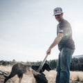 A man in a t-shirt and a Rural Cloth Bull Spurs Icon Hat is shoveling dirt while a black dog stands nearby. They are outdoors in a grassy area with trees and mountains in the background. The sun is shining brightly, creating lens flare.