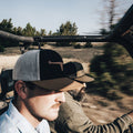 Two men, both wearing Rural Cloth's Bull Spurs Icon hats, are riding in an open-top vehicle in a rural area with trees and dry grass in the background. The motion blur in the photo suggests they are moving quickly. The man in front is driving, and the man in the back is a passenger.