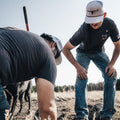 Two men sporting Rural Cloth's Bull Spurs Icon hats and casual attire work outdoors in a field. One is bending over with tools nearby, while the other stands observing. A black dog is also present in the background. The sky is clear with trees visible on the horizon.