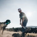 A man wearing the Rural Cloth Bull Spurs Icon Hat and jeans is shoveling dirt outdoors on a sunny day. A large dog stands nearby, looking at the man. Sunlight creates a lens flare effect, and trees are visible in the background.