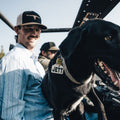 A man wearing a Rural Cloth Bull Spurs Icon hat and a light blue plaid shirt smiles while sitting in an off-road vehicle. A black dog with a tag reading 