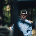 A man wearing a light blue shirt and a Rural Cloth Bull Spurs Icon Hat, distinguished by its longhorn emblem, is standing outdoors. The background is dimly lit and appears to be a forested area. The focus is on the man, while other elements in the foreground are blurred.