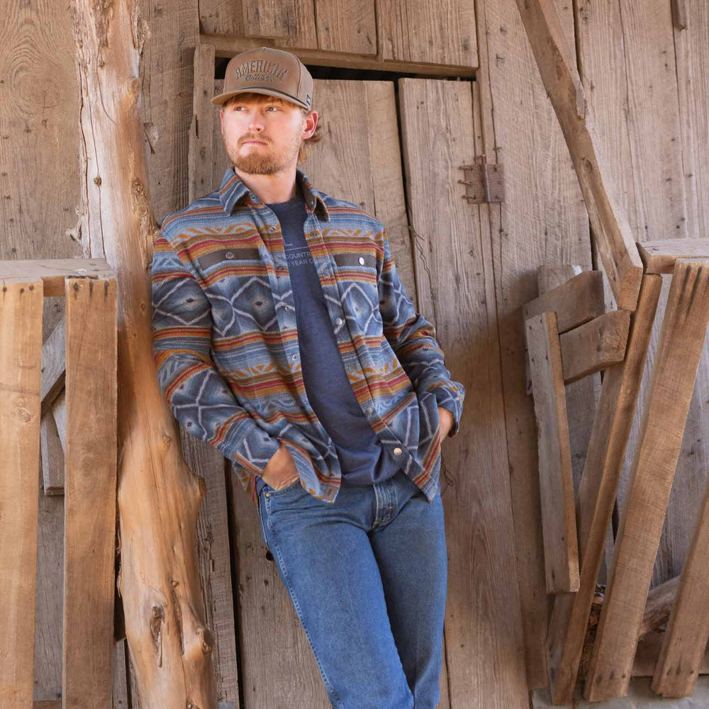 A man with a beard, wearing a patterned button-up shirt and jeans topped by the high-quality Desperado Polar Fleece from Rural Cloth, leans against a wooden barn door with his hands in his pockets. The setting appears rustic, with weathered wood and a rough-hewn tree trunk visible.