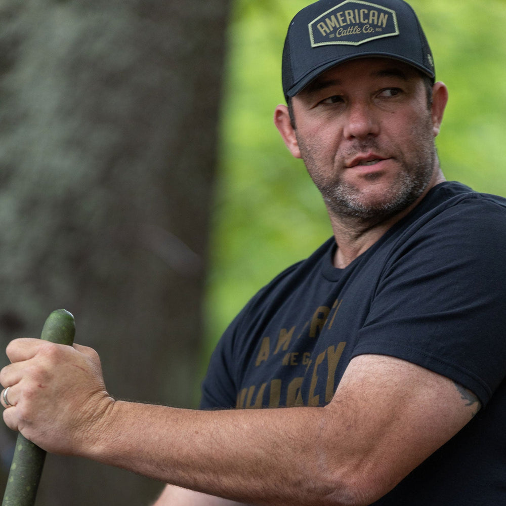 A man sporting a black Diamond Hat from Rural Cloth and a black t-shirt is holding a green pole. The classic snapback style of his cap adds to its appeal. He has a slight smile and a short beard, standing outdoors with a blurred background of greenery and trees.