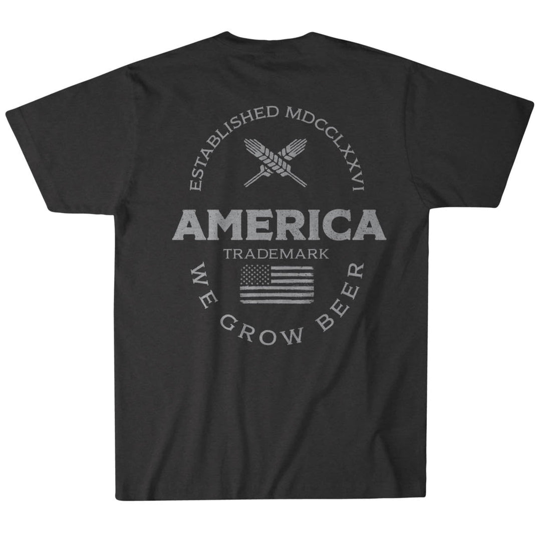 The image displays the back of Rural Cloth's Established Tee, a dark, premium weight shirt. It features crossed wheat stalks with "Established MDCCLXXVI" above, "America Trademark" centered, "We Grow American Beer" below, and includes a flag image.