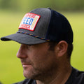 A man is shown in profile wearing the Farmers Feed America Hat by Rural Cloth, characterized by its blue and grey design and featuring a red and white patch with 