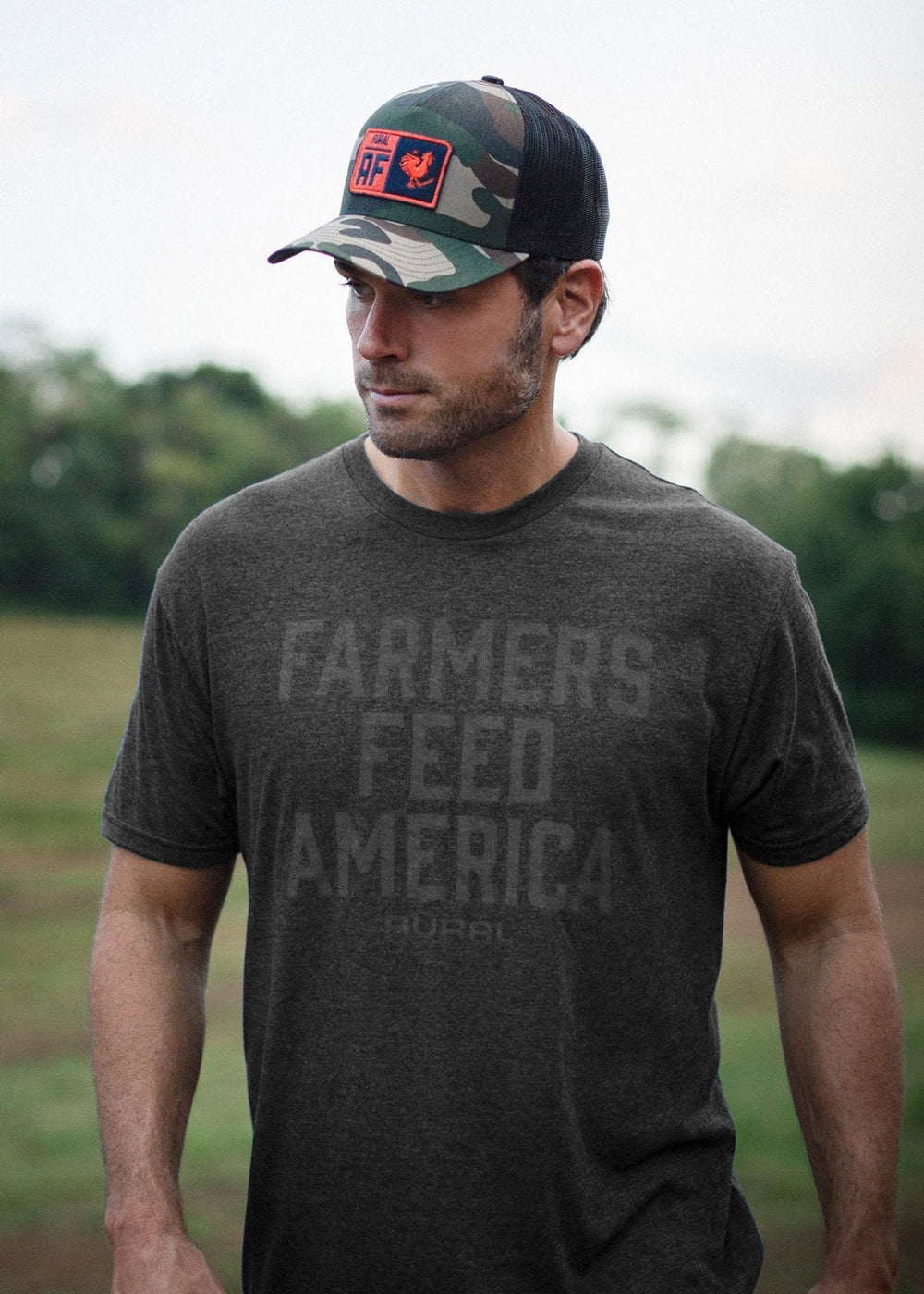 A man wearing Rural Cloth's "Farmers Feed America Tee" made from American-grown cotton and a camo baseball cap stands outdoors. The background is a blurred green field with trees, giving the scene a relaxed, vintage fitted look.