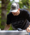 A man with a beard, wearing a cap and a black Rural Cloth Hunt America Tee made of premium weight fabric, is working or examining something in front of him outdoors. The background is blurred, highlighting the lush greenery behind him.