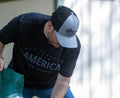 A person wearing a black and grey cap and a Rural Cloth Hunt America Tee bends over while holding a green bag. The background is blurred and features a wall with partial sunlight.