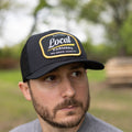 A man with a beard and mustache is wearing the Rural Cloth Local Farmer Hat in black, which features a patch that reads 