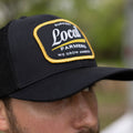 A man is wearing the Rural Cloth Local Farmer Hat-Black. The cap showcases a rectangular patch with the text: 