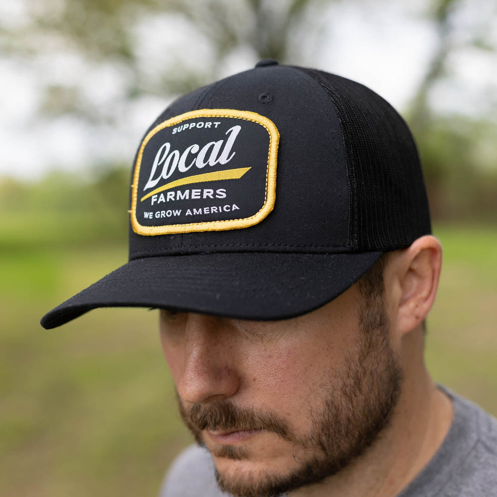 A bearded man, embodying community support, wears the Local Farmer Hat-Black by Rural Cloth. The black baseball cap features a yellow-outlined patch with the message "Support Local Farmers. We Grow America." He stands outdoors, surrounded by blurred greenery.