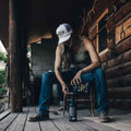 A person with long hair sits on a wooden porch of a rustic log cabin, pouring liquid from a funnel into a container. They are wearing a Rural Cloth Longhorn Hat, tank top, jeans, and slippers. Various tools and objects are scattered around, suggesting a hands-on task or repair work in their ranching lifestyle.