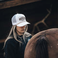 A person with long hair dons a white Longhorn Hat from Rural Cloth and a black shirt. Embracing the ranching lifestyle, the individual stands next to a brown horse, its back and tail visible in the foreground.