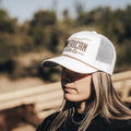 A person with long hair wearing a white Longhorn Hat from Rural Cloth stands outside. The background features blurred vegetation and what appears to be a wooden structure, evoking a ranching lifestyle. The person is looking slightly downward.