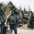 A man with a beard, wearing a Longhorn Hat from Rural Cloth, sunglasses, a hoodie, and jeans, walks through a grassy area next to an off-road vehicle. The background features trees and a landscape with dry grass and rolling hills, capturing the essence of the ranching lifestyle.