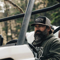 A bearded man wearing a Longhorn Hat by Rural Cloth sits in a vehicle, looking to his right. He is dressed in a hooded sweatshirt with a deer logo on the front. The vehicle structure and outdoors with trees are visible in the background, evoking a ranching lifestyle.